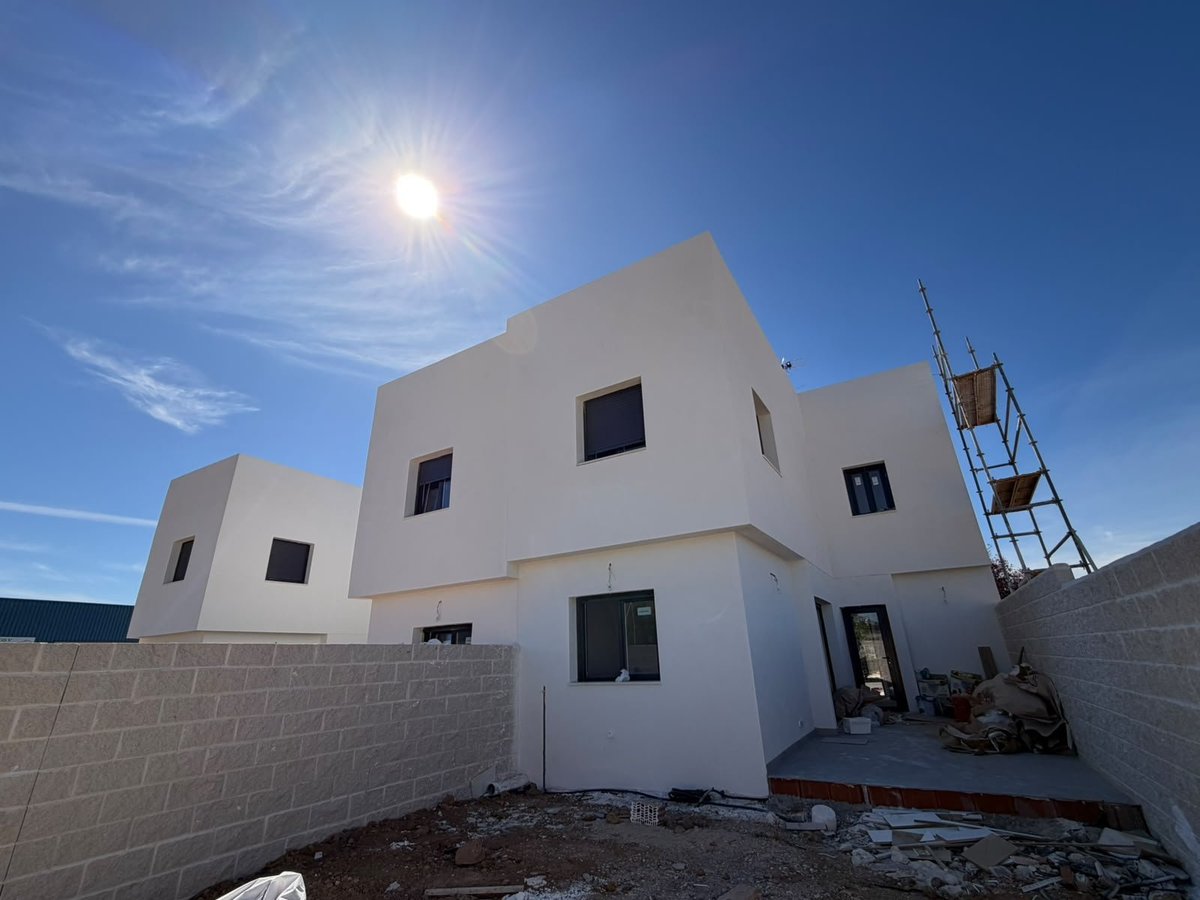Bloque de chalets adosados de Grama Homes en construcción con fachada blanca, ventanas cuadradas y andamios bajo un cielo azul con sol