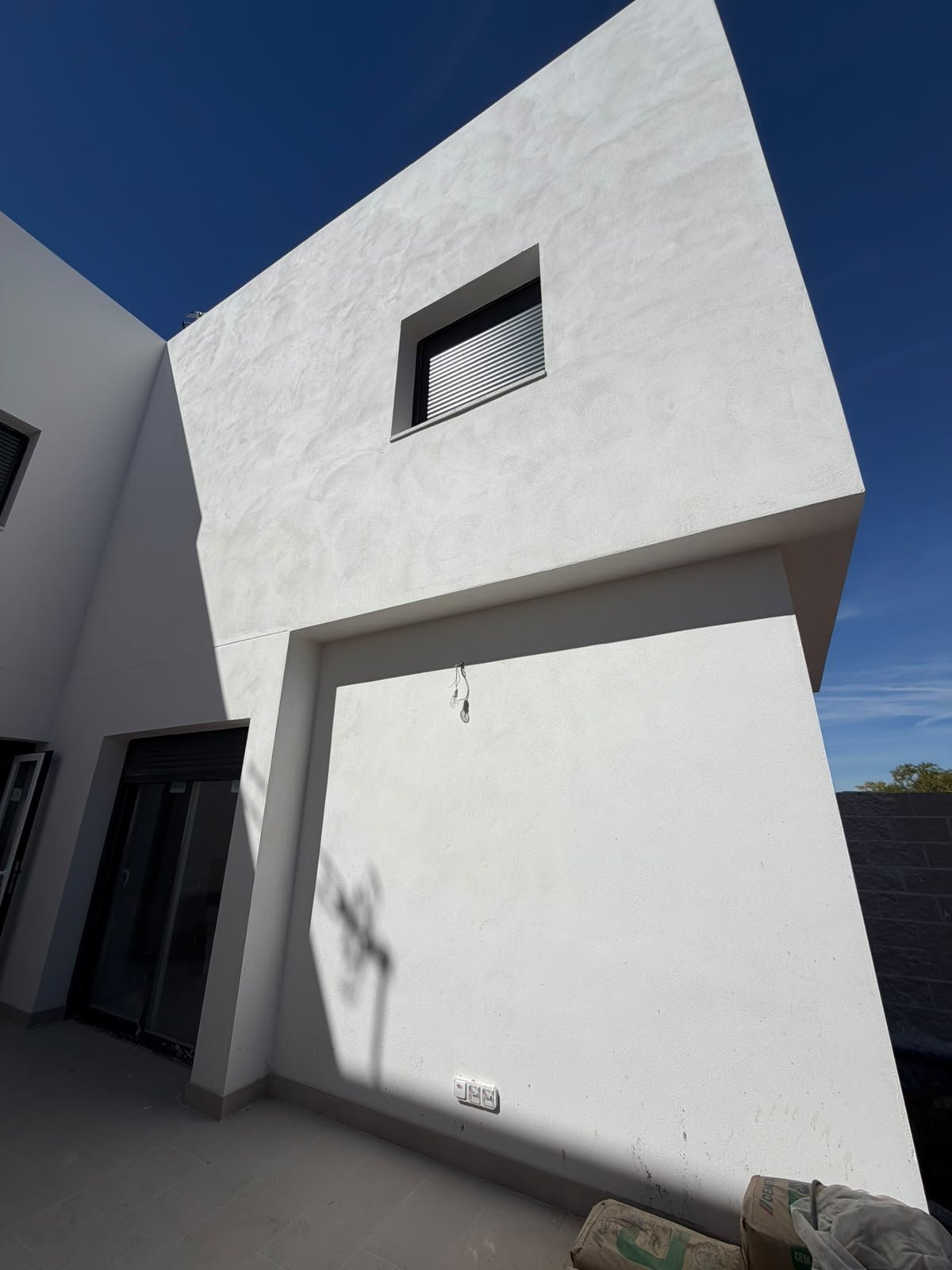 Detalle arquitectónico de uno de los chalets adosados de Grama Homes con fachada blanca cúbica, una ventana superior y entrada oscura bajo un cielo azul