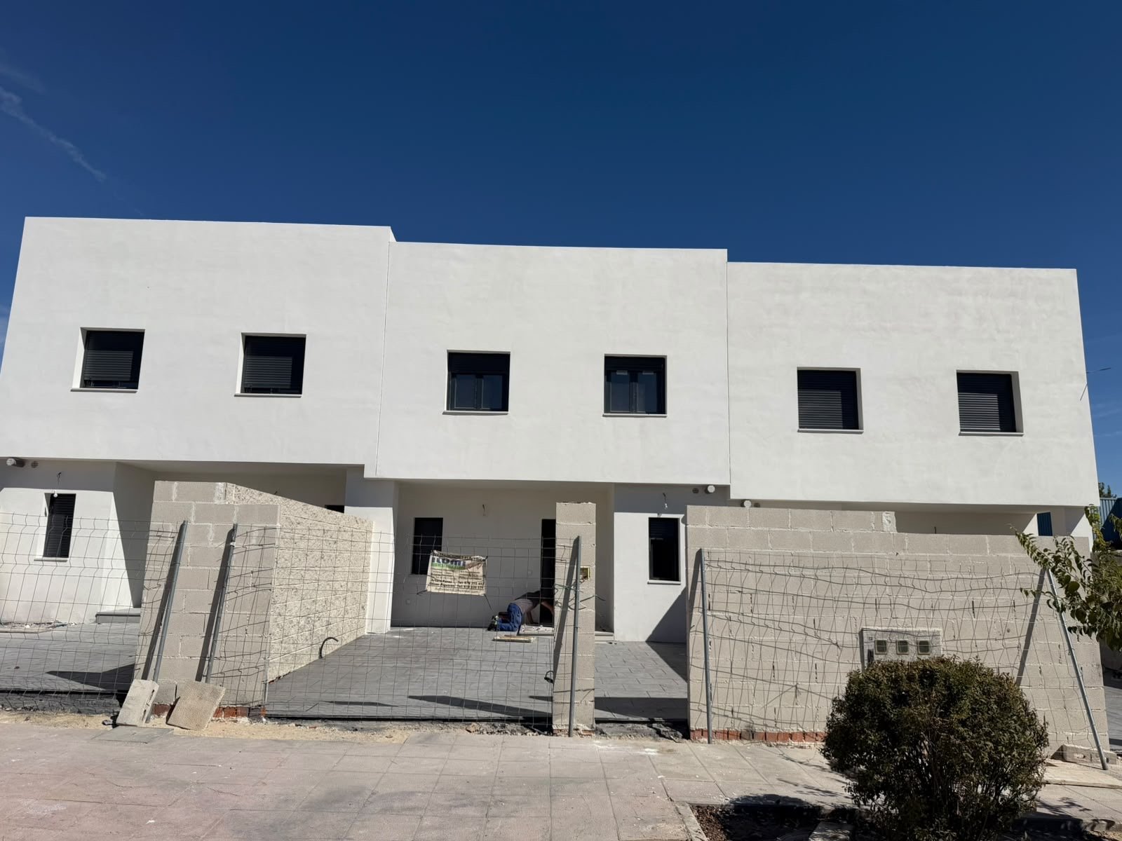 Vista exterior de los chalets adosados de Grama Homes en Valmojado con fachada blanca cúbica, pequeñas ventanas cuadradas y valla perimetral de obra bajo un cielo azul